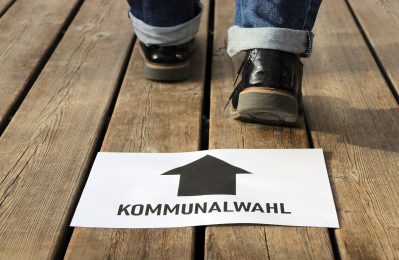 Zettel mit Pfeil und "Kommunalwahl" - über dem Zettel sind Füße, die gehen zu sehen